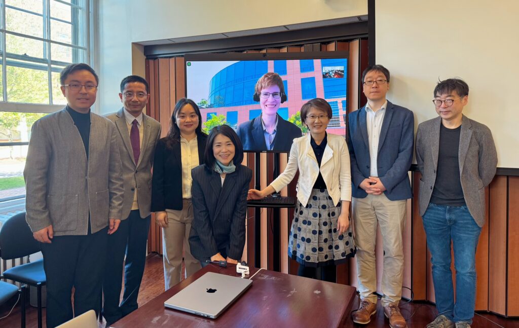 7 people in a classroom pose in front of a TV screen which shows an 8th person attending virtually