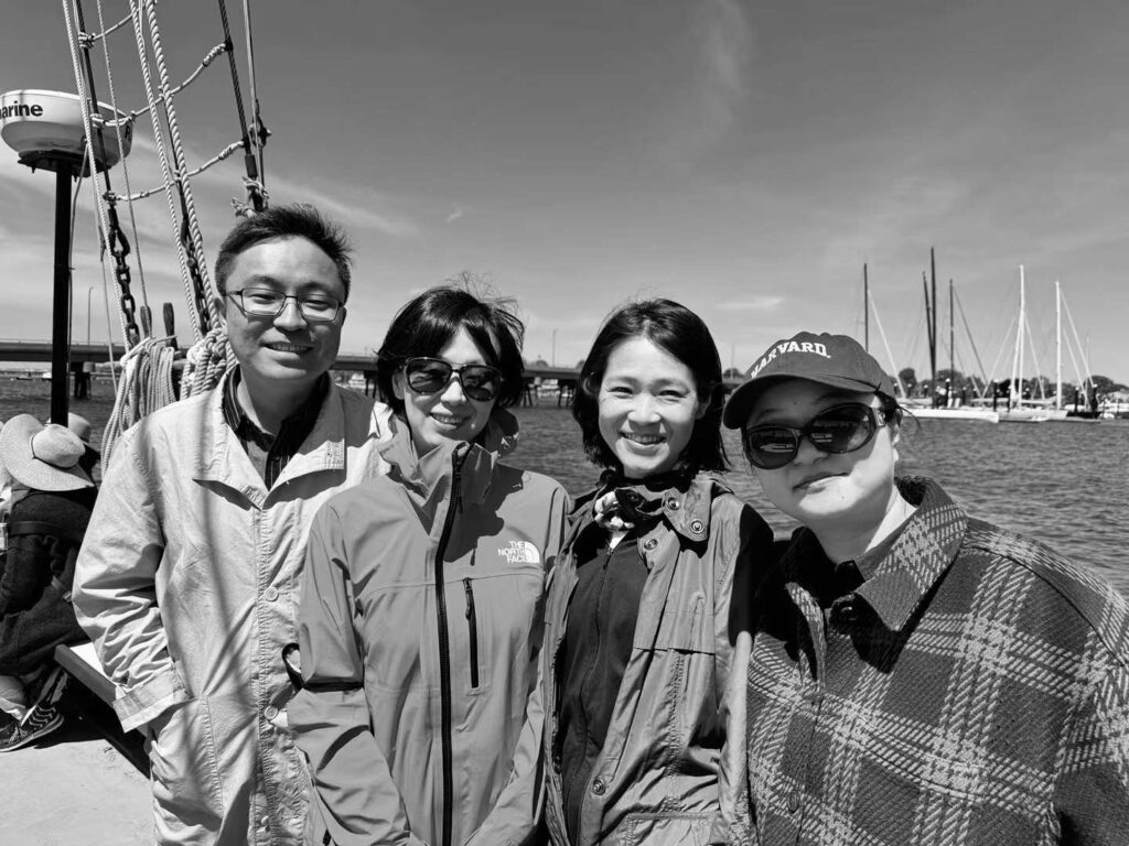 Four people stand on a sailboat with water and other boats in the background